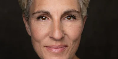 Close-up portrait of a middle-aged person with short, light hair, brown eyes, and a neutral expression against a dark background.