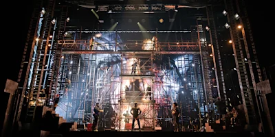 A stage set with metal scaffolding, dramatic lighting, and performers in silhouette during a theatrical production.