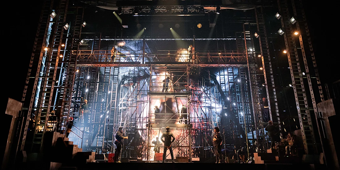 A stage set with metal scaffolding, dramatic lighting, and performers in silhouette during a theatrical production.