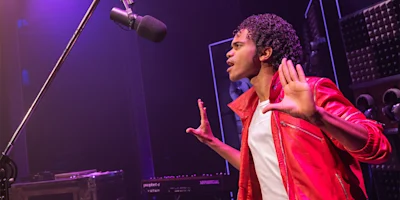 A young man in a red jacket and white shirt stands in front of a microphone with his hands raised, appearing to sing or perform on stage under purple lighting.