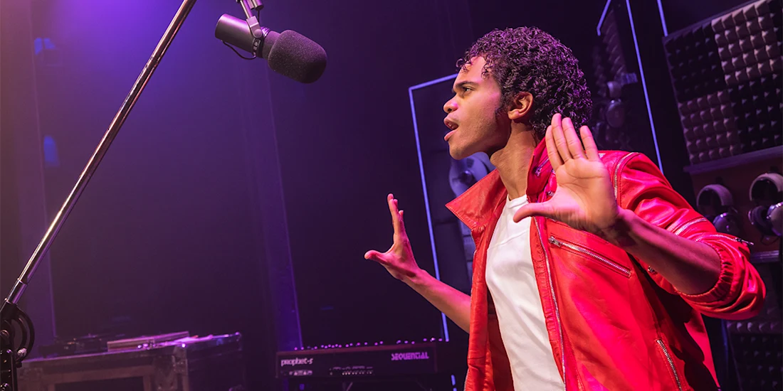 A young man in a red jacket and white shirt stands in front of a microphone with his hands raised, appearing to sing or perform on stage under purple lighting.