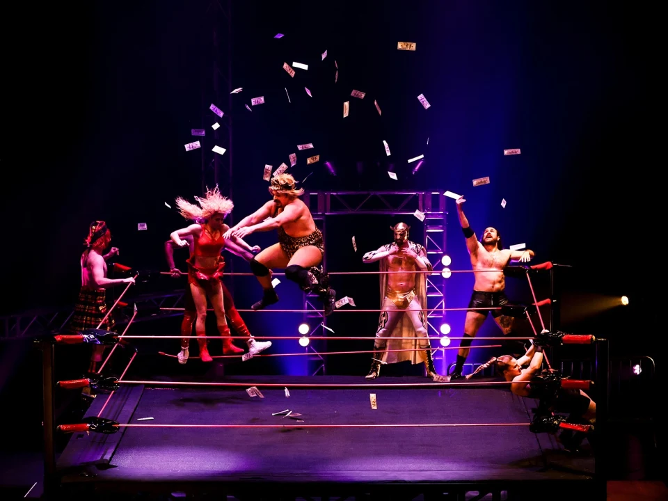 A group of wrestlers performs in a ring with money flying through the air amid bright stage lighting.