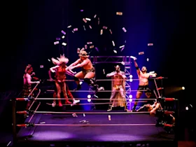 A group of wrestlers performs in a ring with money flying through the air amid bright stage lighting.