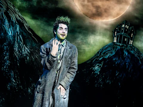 A person in theatrical makeup and tattered clothing performs on stage with a painted backdrop of hills, a full moon, and a house.