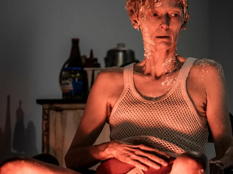 A person wearing a mesh tank top, with white substance on their face, neck, and shoulders, sitting in dim light.