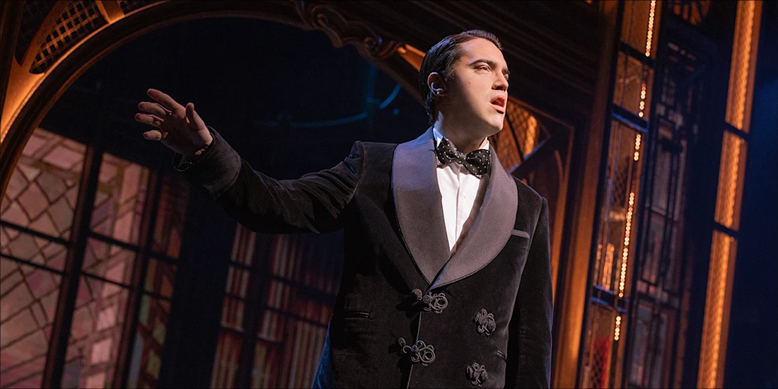 A performer in a black suit and bow tie stands onstage with one arm raised, set against an ornate backdrop with warm lighting.