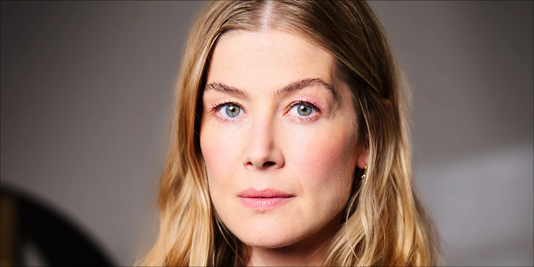 A woman with long, wavy blonde hair and light eyes looks directly at the camera with a neutral expression, against a softly blurred background.