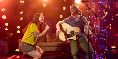 Woman singing into a microphone and man playing guitar on stage, both smiling, with colorful lights and a sofa in the background.