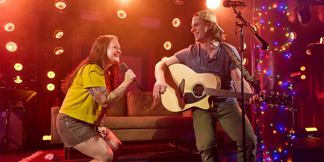 Woman singing into a microphone and man playing guitar on stage, both smiling, with colorful lights and a sofa in the background.