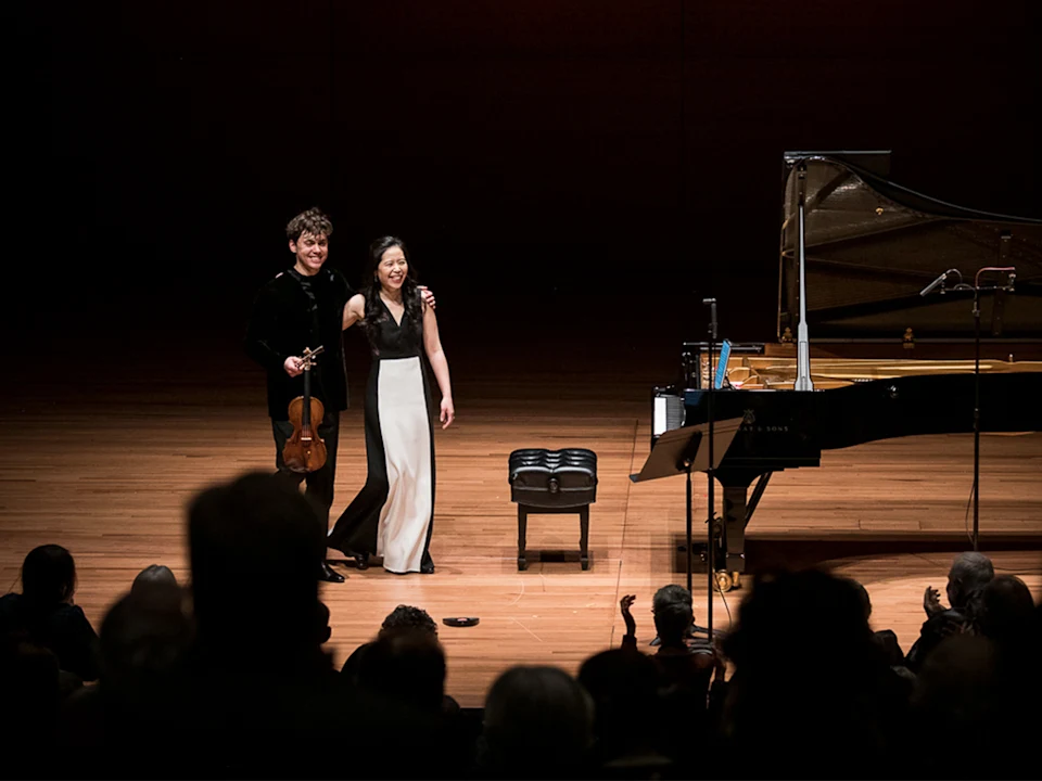 Two musicians stand on stage, one holding a violin, next to a grand piano, facing an applauding audience in a concert hall.