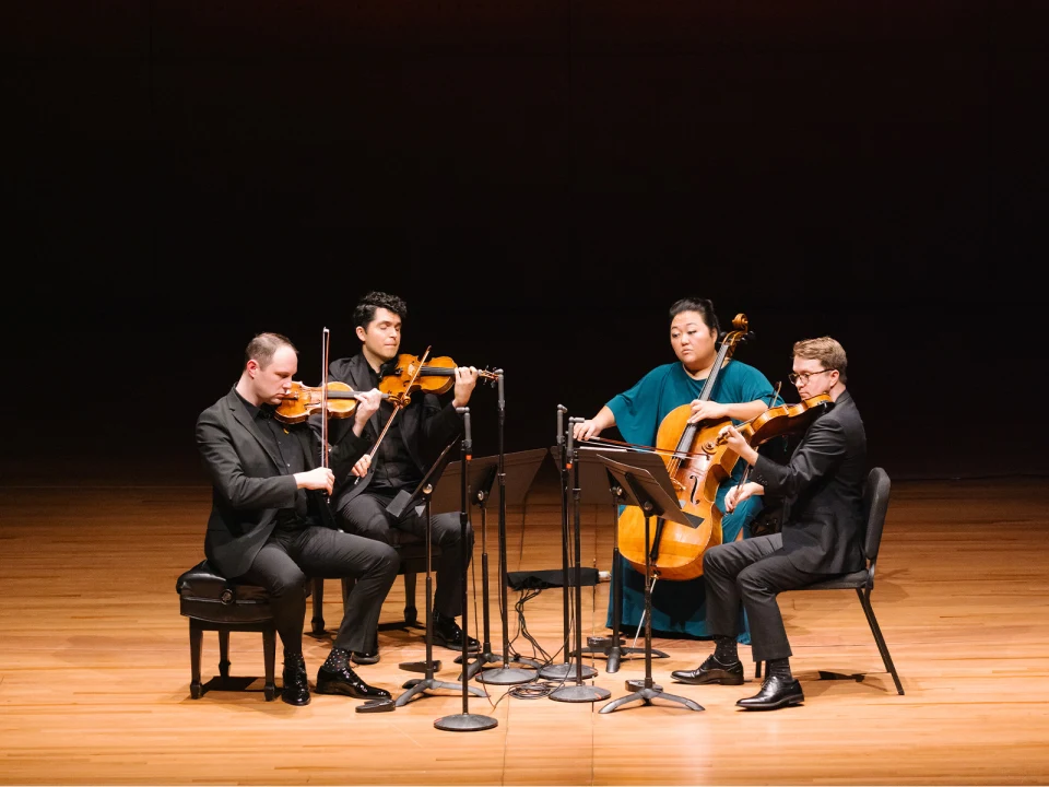 A string quartet performs on stage, with two violinists, a violist, and a cellist, all seated and focused on their instruments. Musical stands with sheet music are placed in front of them.