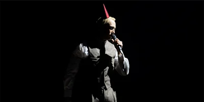 A person wearing a gray vest, white shirt, and a red cone hat stands in a spotlight, holding a microphone against a dark background.