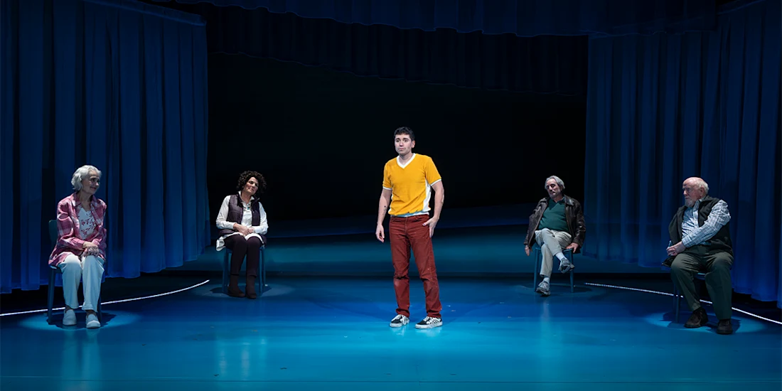 Five people are on stage with blue curtains; one stands center in a yellow shirt and red pants, while the other four are seated and spaced apart, looking toward the center.
