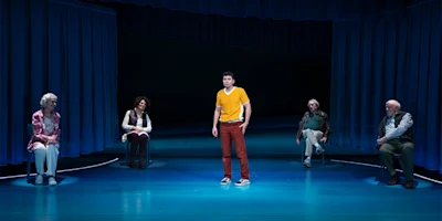 Five people are on stage with blue curtains; one stands center in a yellow shirt and red pants, while the other four are seated and spaced apart, looking toward the center.