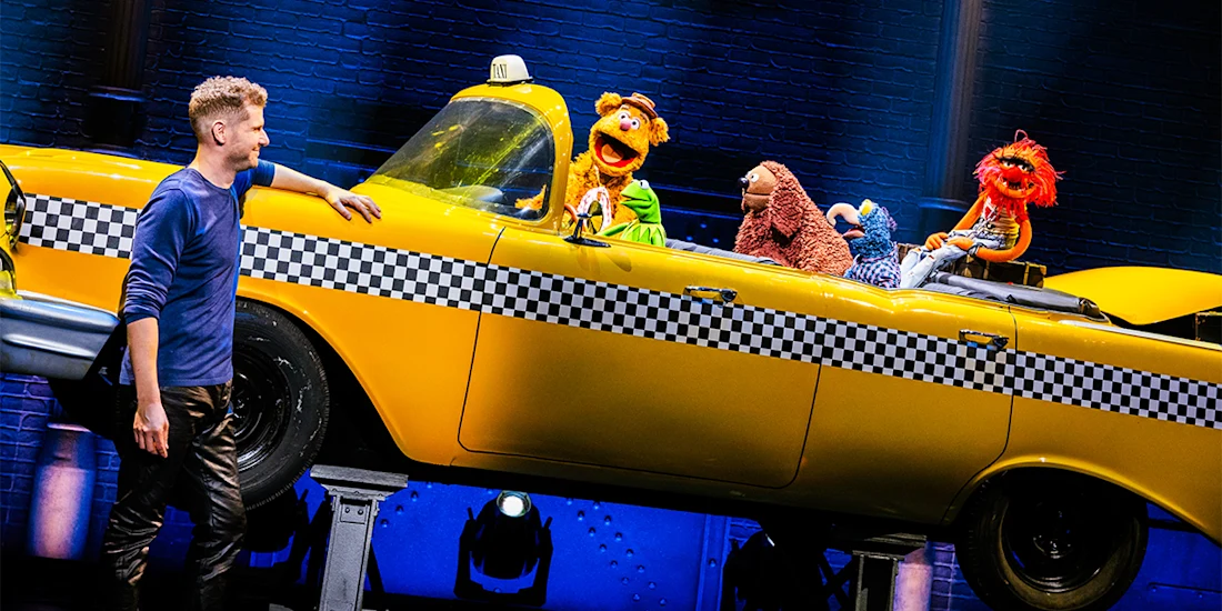 A man stands next to a yellow taxi cab on stage with five Muppet characters sitting inside, under theater lighting.