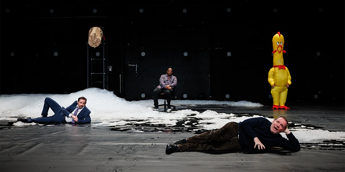 Three men pose on a stage with artificial snow and a ladder; an inflatable chicken stands upright in the background.