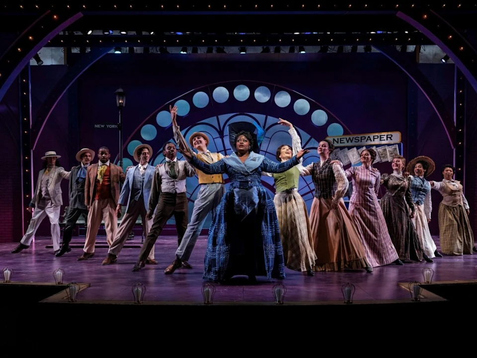 A group of actors in period costumes perform a musical number on stage, with a lit "NEWSPAPER" sign and an arched window backdrop behind them.