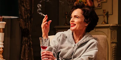 A woman in a gray hoodie sits indoors, smiling while holding a glass of red wine in one hand and a lit cigarette in the other.