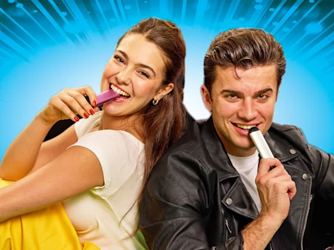 A woman and a man sitting back to back, smiling, each holding and vaping from a colorful vape device against a bright blue background.