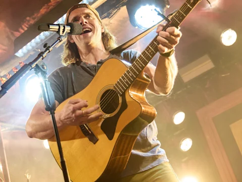 A person wearing a backwards cap plays an acoustic guitar and sings into a microphone on stage under bright lights.