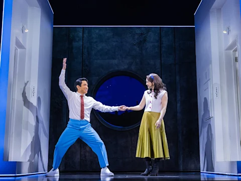 A man in blue pants and a white shirt enthusiastically holds hands with a woman in a white top and yellow skirt on a stage with blue-lit doors and a circular window backdrop.