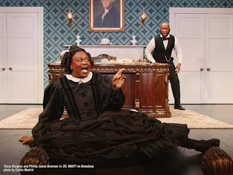 A person in a black dress sits on the floor in front of a desk, gesturing animatedly, while another person in formal attire stands behind the desk looking surprised.