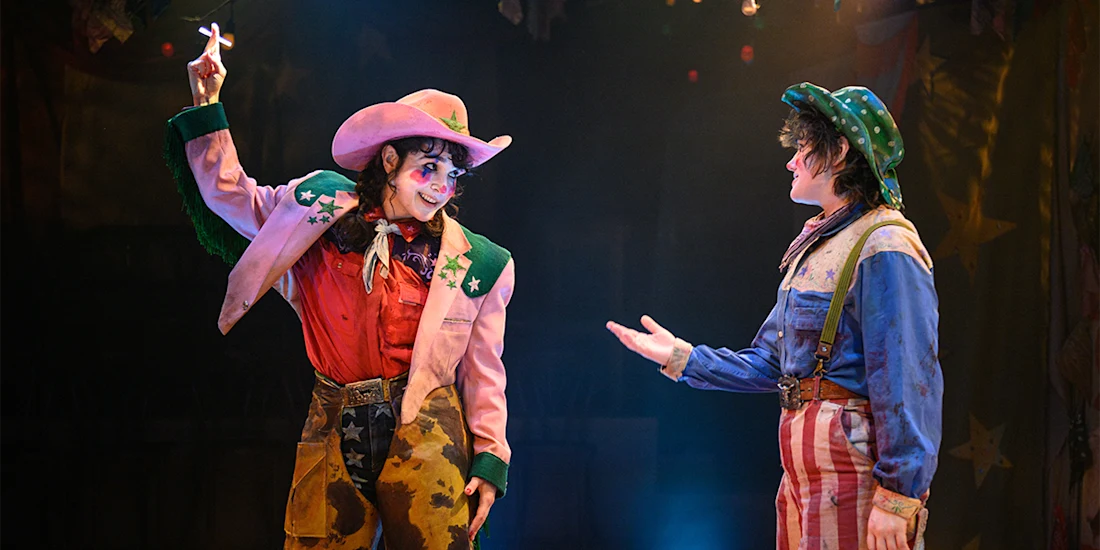 Two performers dressed as colorful clowns in Western-style costumes interact on stage under theatrical lighting.