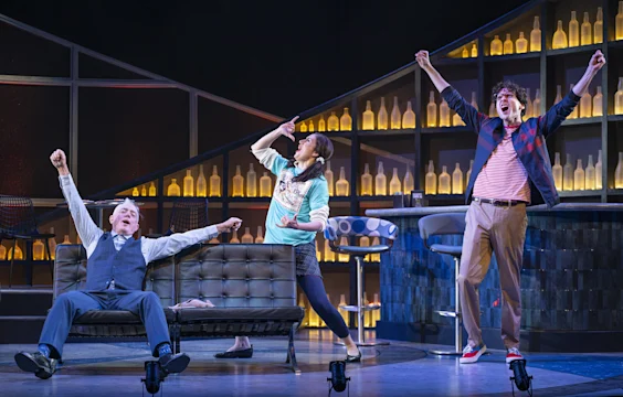 Three actors on a stage set resembling a bar, with one man seated and two people standing, all raising their arms in expressive poses. Shelves of bottles are visible in the background.