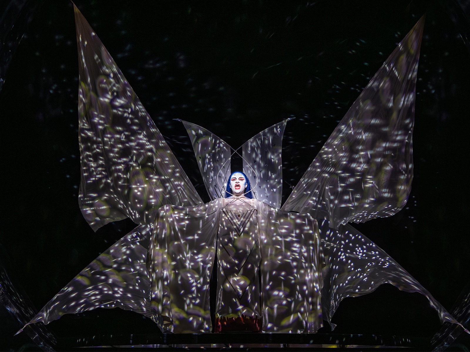 A performer stands center stage, illuminated in a dramatic costume with large, wing-like fabric panels and star-like light projections covering the scene.