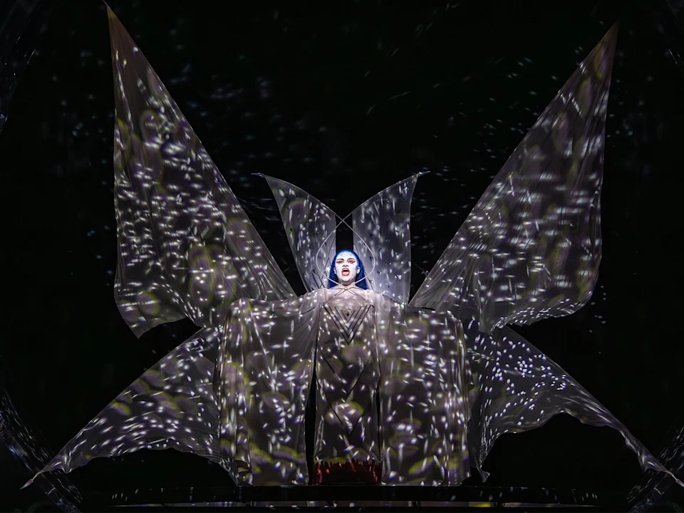 A performer stands center stage, illuminated in a dramatic costume with large, wing-like fabric panels and star-like light projections covering the scene.