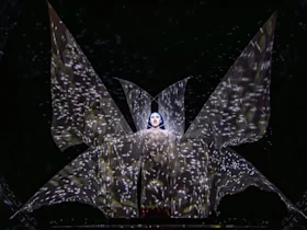 A performer stands center stage, illuminated in a dramatic costume with large, wing-like fabric panels and star-like light projections covering the scene.