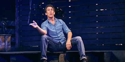 A man in a blue shirt and jeans sits on a stage bench, gesturing with his right hand and speaking, with a dark wooden backdrop behind him.