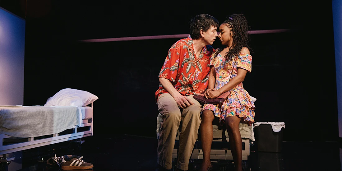 Two people sit closely on a bed, gazing at each other. The scene is set on a dimly lit stage with minimal props, including a pair of shoes on the floor and a neatly made bed.