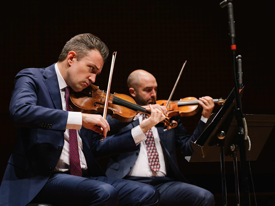 Two men in suits play violins on stage, seated next to each other with sheet music and a microphone stand visible.