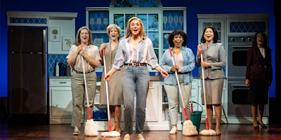 Six women stand on stage in a kitchen set, five holding mops and smiling, with one woman singing at the front and center.