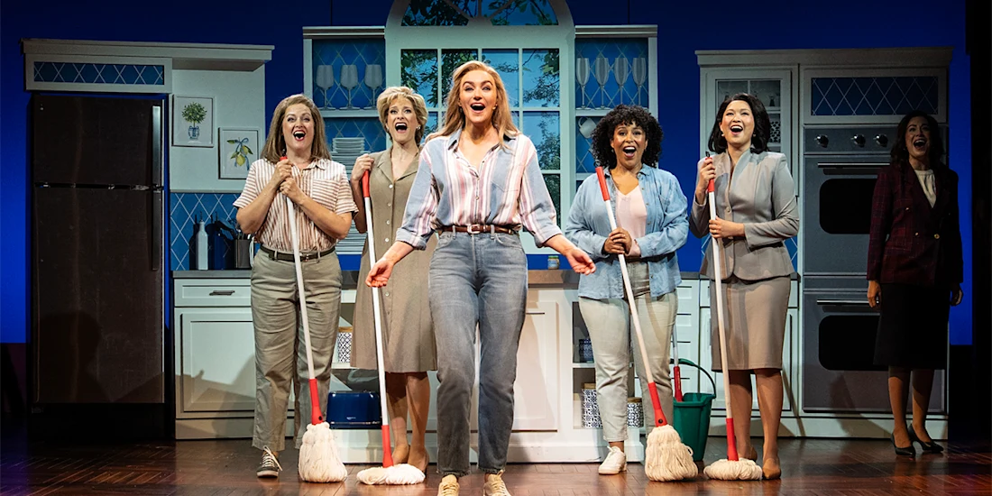 Six women stand on stage in a kitchen set, five holding mops and smiling, with one woman singing at the front and center.