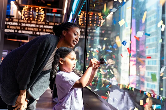 An adult and child look through a glass window covered in colorful confetti, with bright lights and reflections visible on the other side.