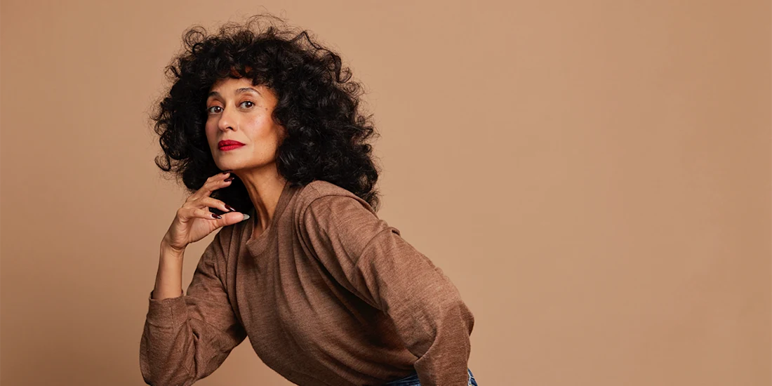 A person with curly dark hair, wearing a brown long-sleeve shirt and red lipstick, poses against a plain beige background.