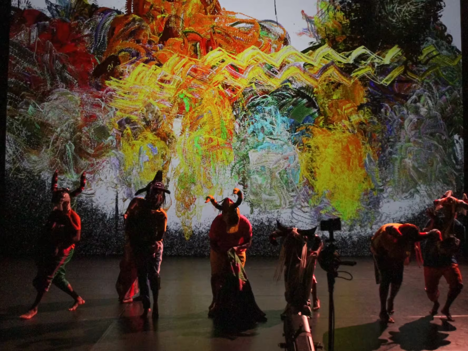 Performers in colorful costumes and masks dance on stage in front of a vibrant, abstract digital backdrop. A camera is visible in the foreground.