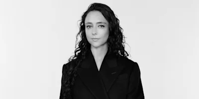 A person with long, wavy dark hair wearing a dark blazer stands against a plain, light background, looking directly at the camera with a neutral expression.