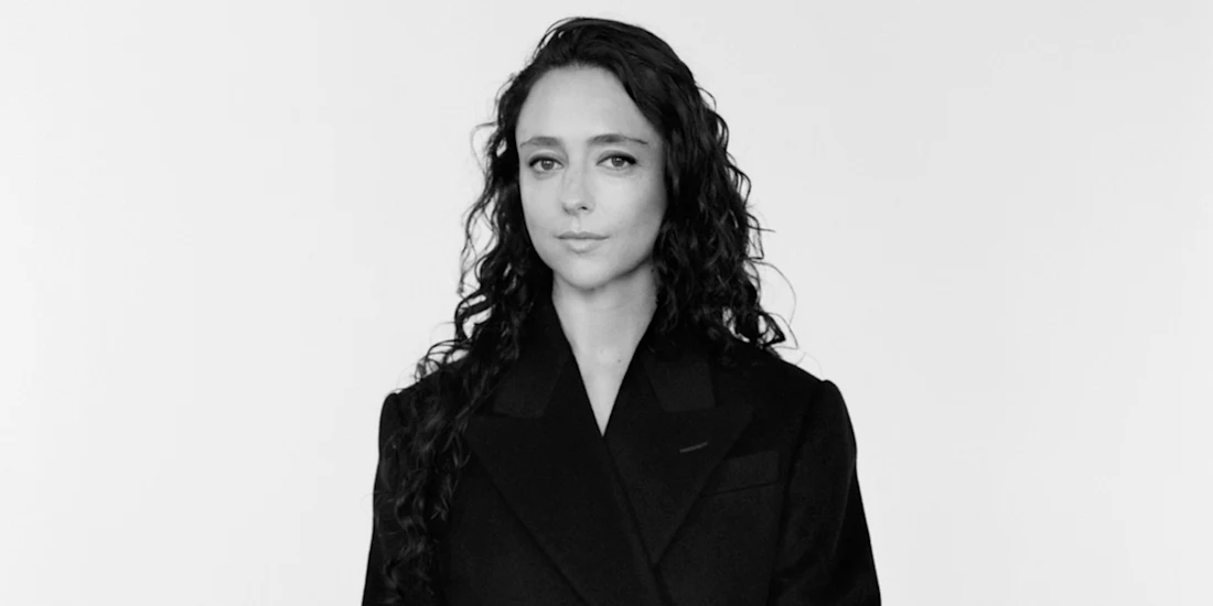 A person with long, wavy dark hair wearing a dark blazer stands against a plain, light background, looking directly at the camera with a neutral expression.