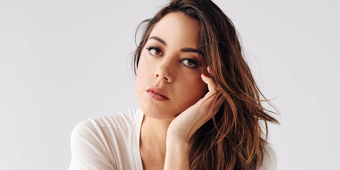 A woman with long brown hair and a neutral expression rests her face on her hand, looking directly at the camera against a plain light background.