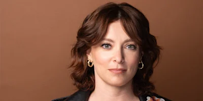 A woman with shoulder-length brown hair, wearing gold hoop earrings and a black jacket, poses in front of a plain brown background.