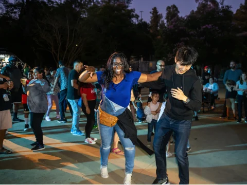 Two people smile and dance together in an outdoor nighttime gathering with a crowd of people in the background.