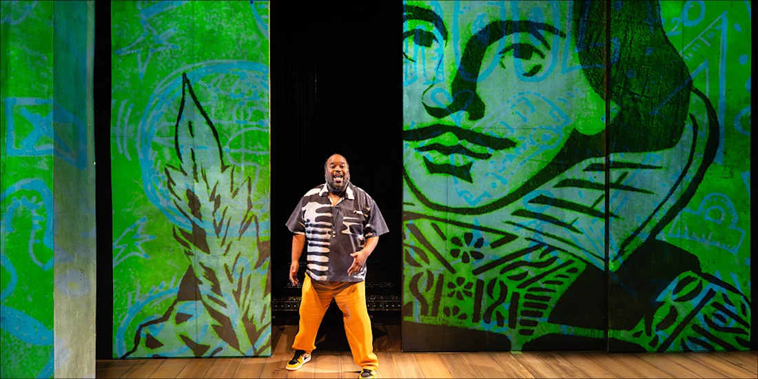 A man stands center stage in front of green panels featuring a large, stylized illustration of William Shakespeare’s face.