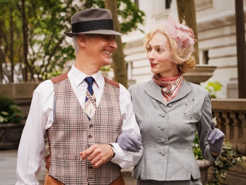 A man and woman in vintage attire walk arm in arm, smiling at each other outside near trees and a stone railing.