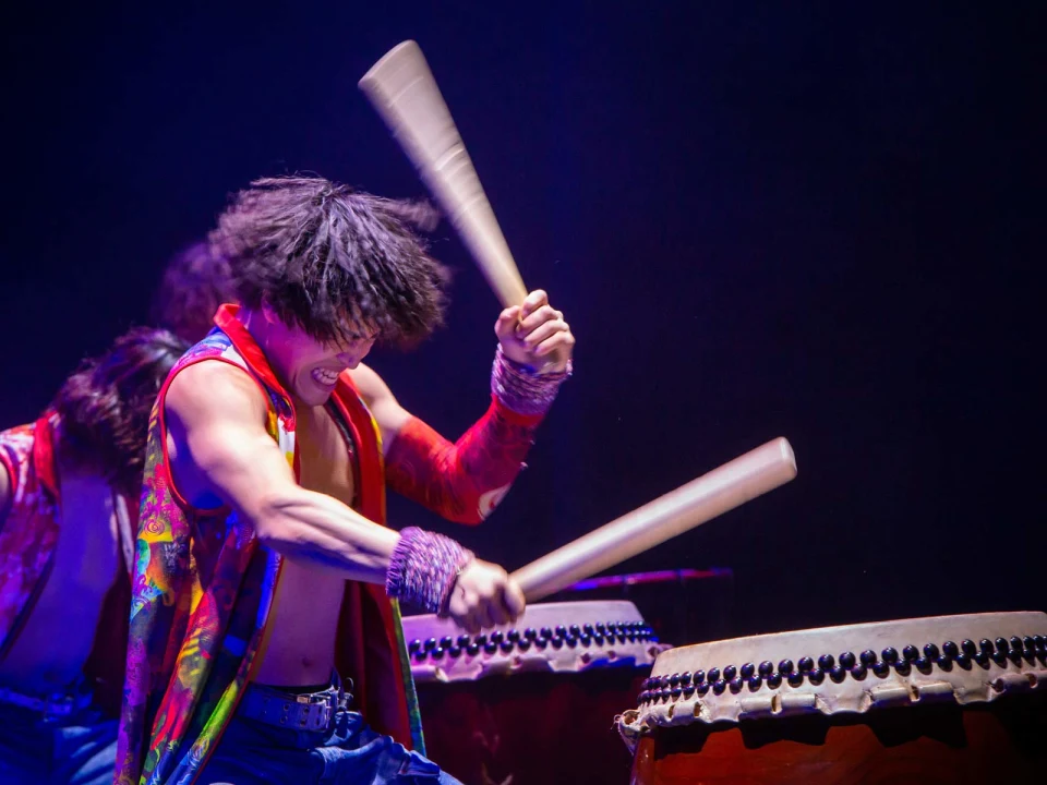 A performer energetically playing a traditional drum with wooden sticks on a dimly lit stage.