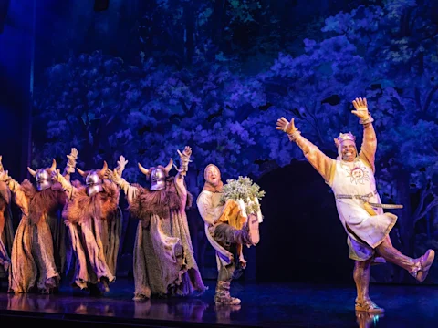 Actors on stage in medieval costumes perform energetically; one holds greenery, others wear horned helmets, and the background depicts a forest scene.