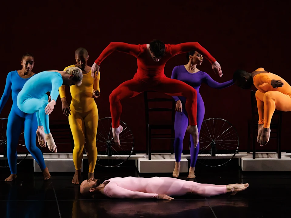 Six dancers in brightly colored bodysuits perform dynamic poses; one lies on the floor while others leap or balance on chairs against a dark background.