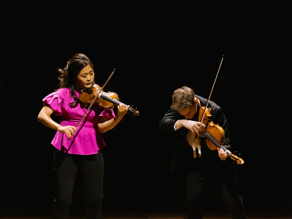 Two violinists perform on stage; a woman in a pink blouse plays a violin on the left, while a man in a black suit bends forward while playing a violin on the right.